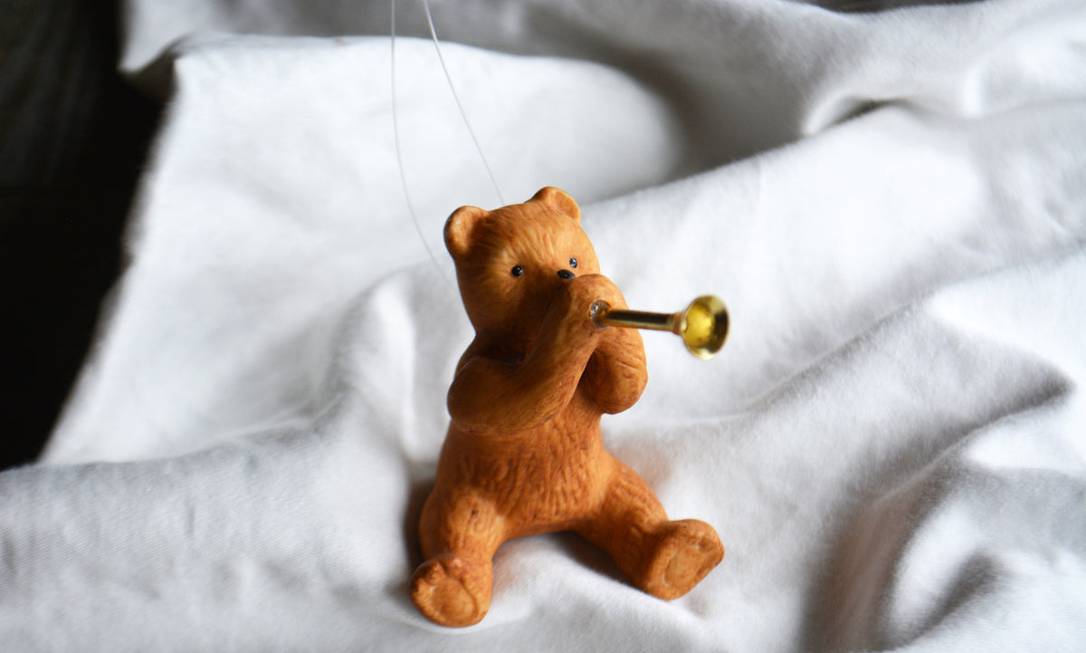 A small wooden teddy bear ornament sitting and playing a gold trumpet, hanging against a draped white fabric background.