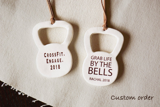 Two white kettlebell-shaped hanging ornaments with brown hanging strings, engraved with custom text, displayed on a textured beige fabric background.