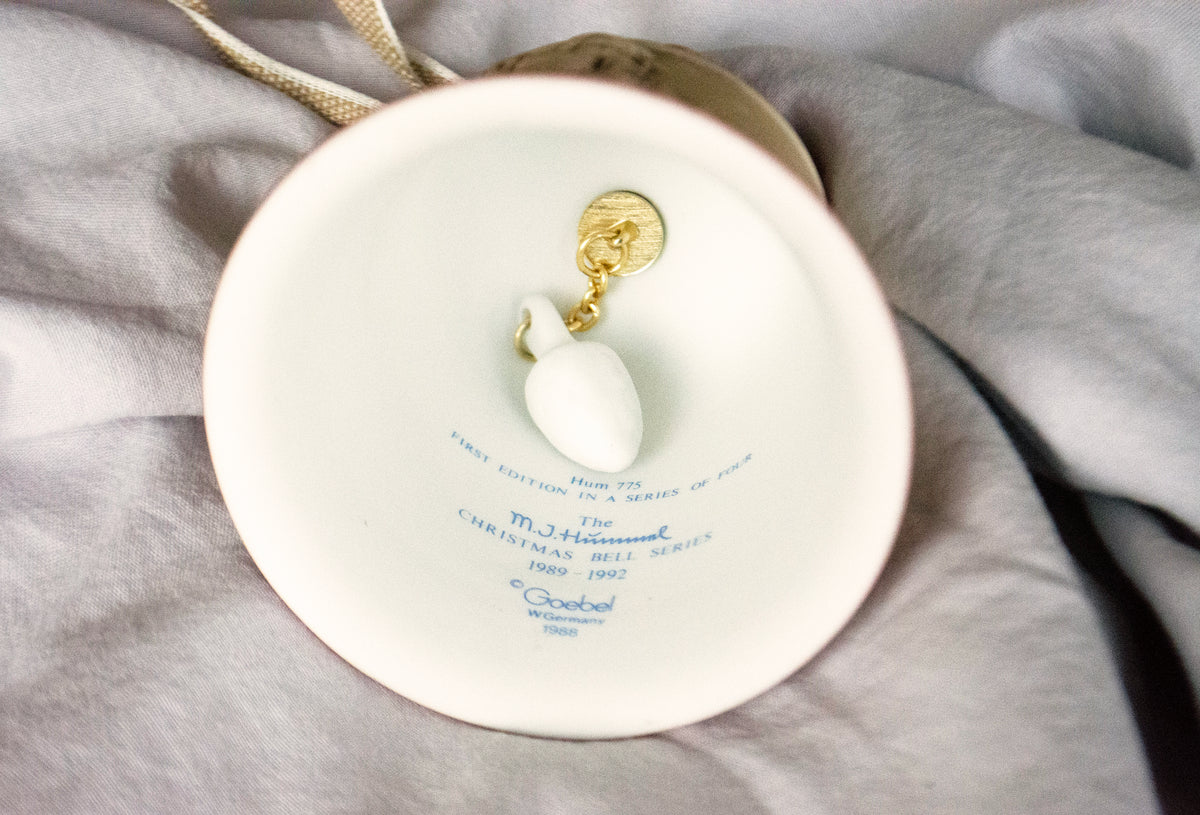 A close-up of a white ceramic ornament holder or display plate with a small white ornament and gold hanging loop, set against soft gray fabric.