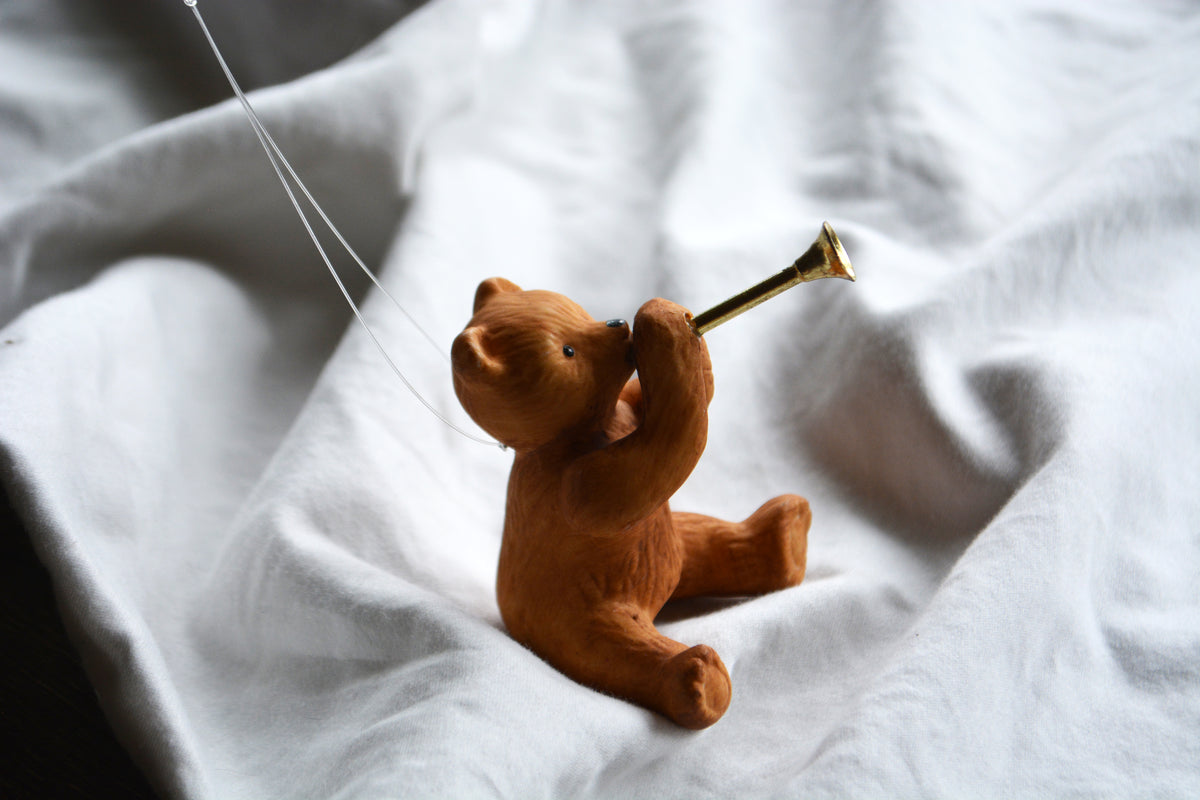 A small brown teddy bear figurine sitting on rumpled white fabric, holding a gold trumpet-like horn.