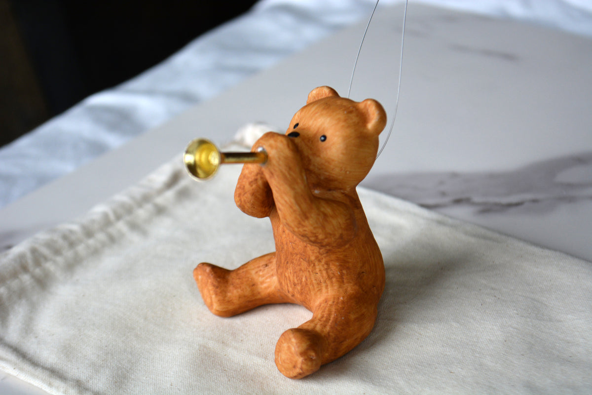 A small carved wooden bear figurine sitting and playing a brass instrument, placed on a light fabric surface with a blurred indoor background.