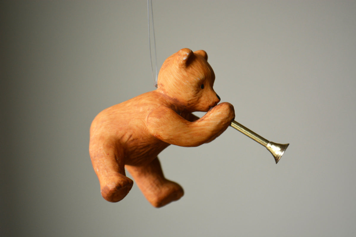 A small wooden bear ornament or figurine holding a brass trumpet, suspended against a plain light gray background.