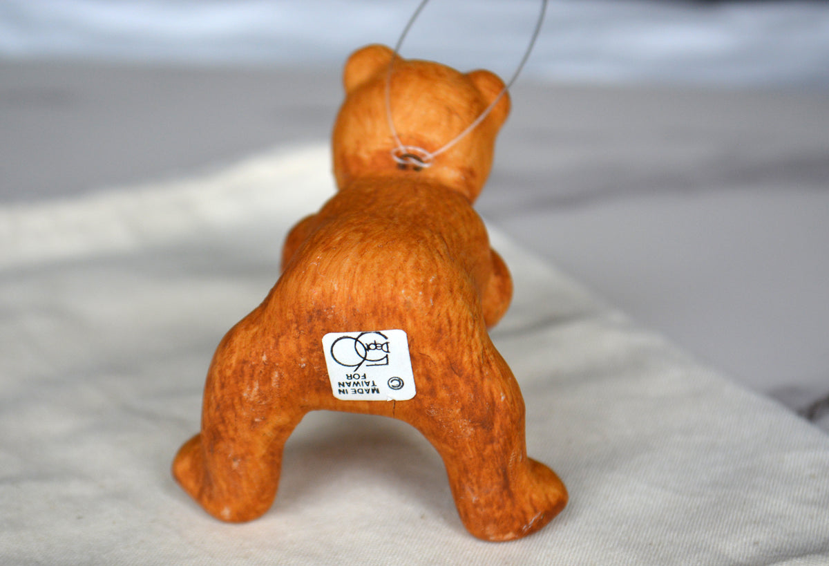 An orange-brown ceramic or clay bear figurine shown from the back, standing on a light fabric surface. A small white sticker with a cartoon bear logo is attached near the base.