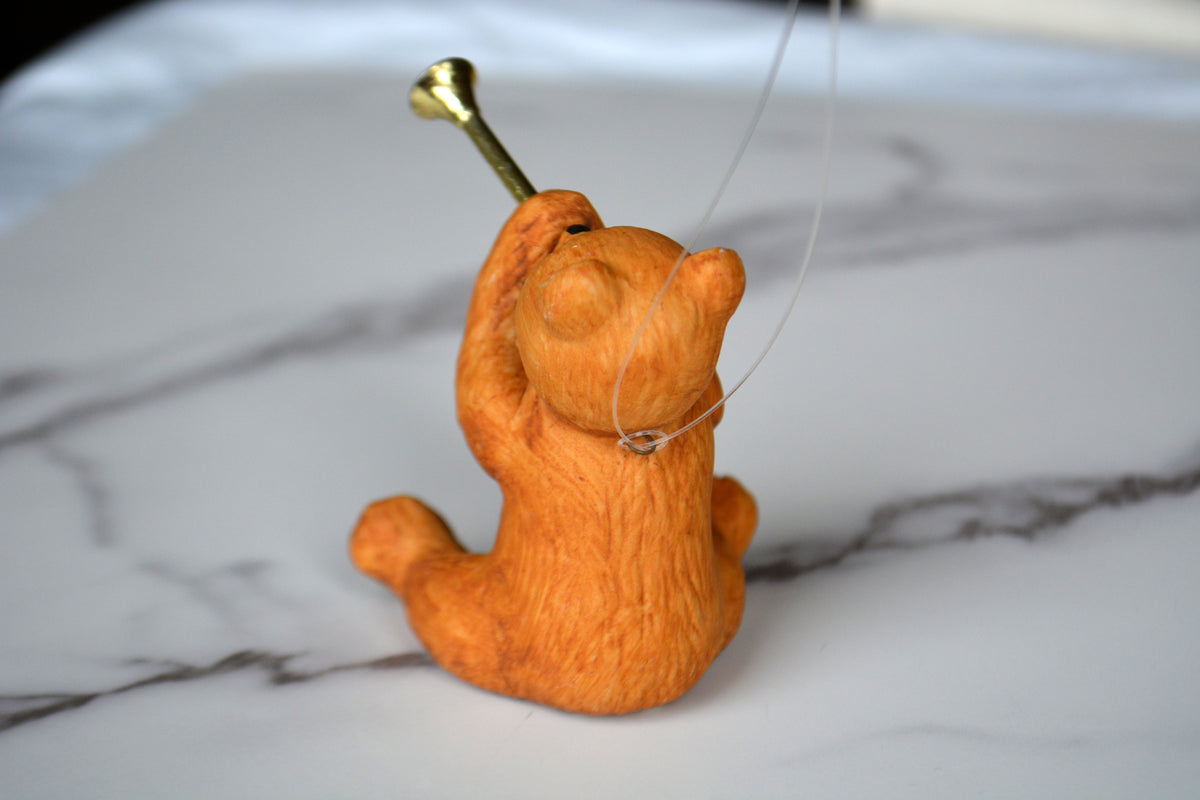 An orange cat figurine sitting on a white marble surface, holding a small gold-colored nail vertically in front of its face, with a thin clear string attached.