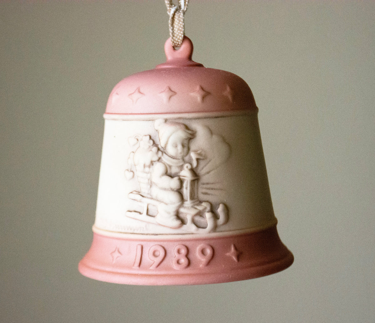 A vintage 1989 Christmas bell ornament featuring a pink and cream-colored ceramic design with a relief of a child and a sleigh, hanging against a neutral gray background.