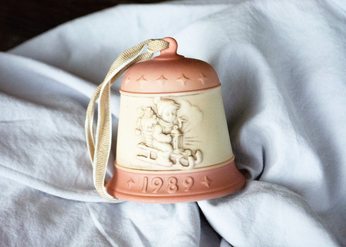 A vintage ceramic Christmas bell ornament with a cream-colored center and terracotta-colored top and bottom, featuring an embossed figure and the year '1989', hanging from a cream ribbon against a draped light gray fabric background.