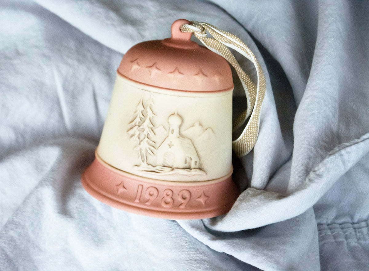 A vintage ceramic Christmas bell ornament with a terracotta-colored top and bottom, and a cream-colored center featuring an embossed winter scene and the year '1989'. It has a beige woven hanging loop and is resting on draped light gray fabric.