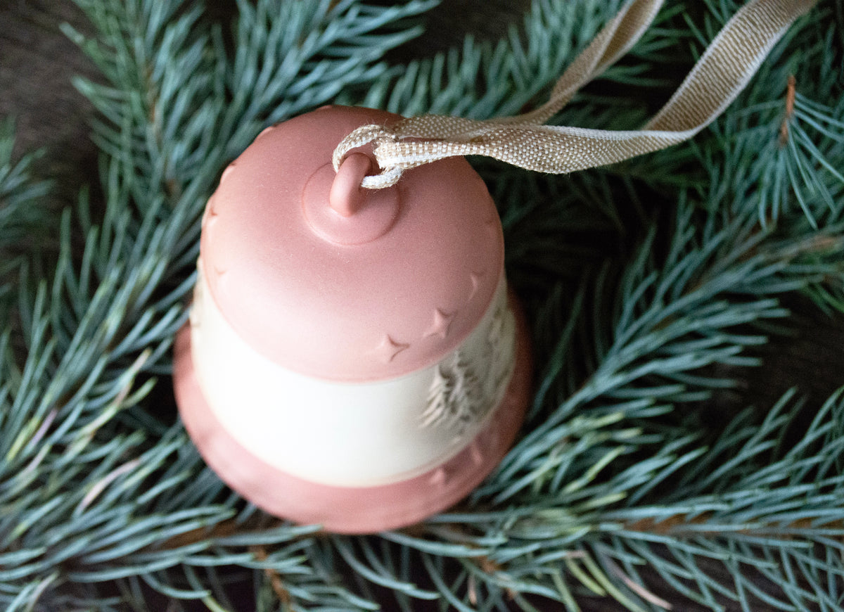 A small decorative bell-shaped Christmas ornament in dusty pink and white, hanging from a beige ribbon, nestled among green pine branches.