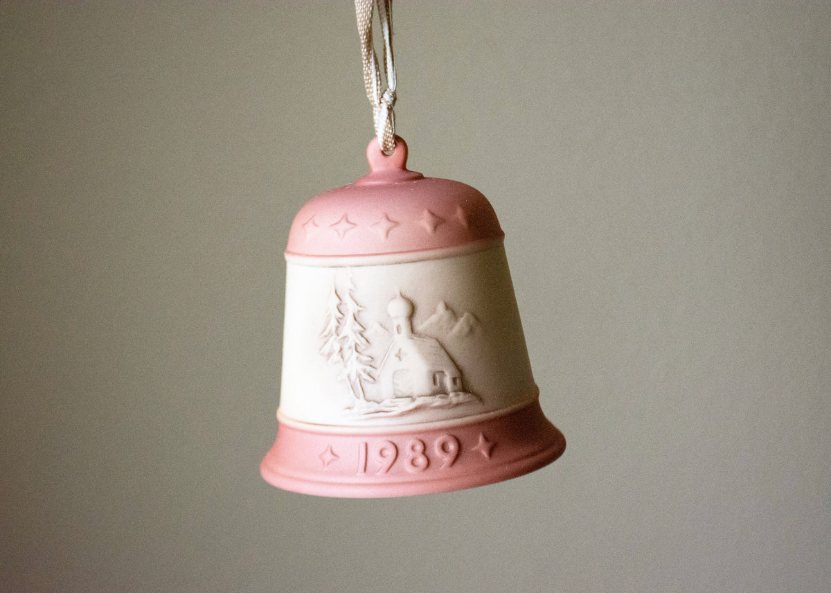 A vintage 1989 ceramic Christmas bell ornament in pink and cream, featuring an embossed winter scene and a hanging loop, shown against a plain gray background.