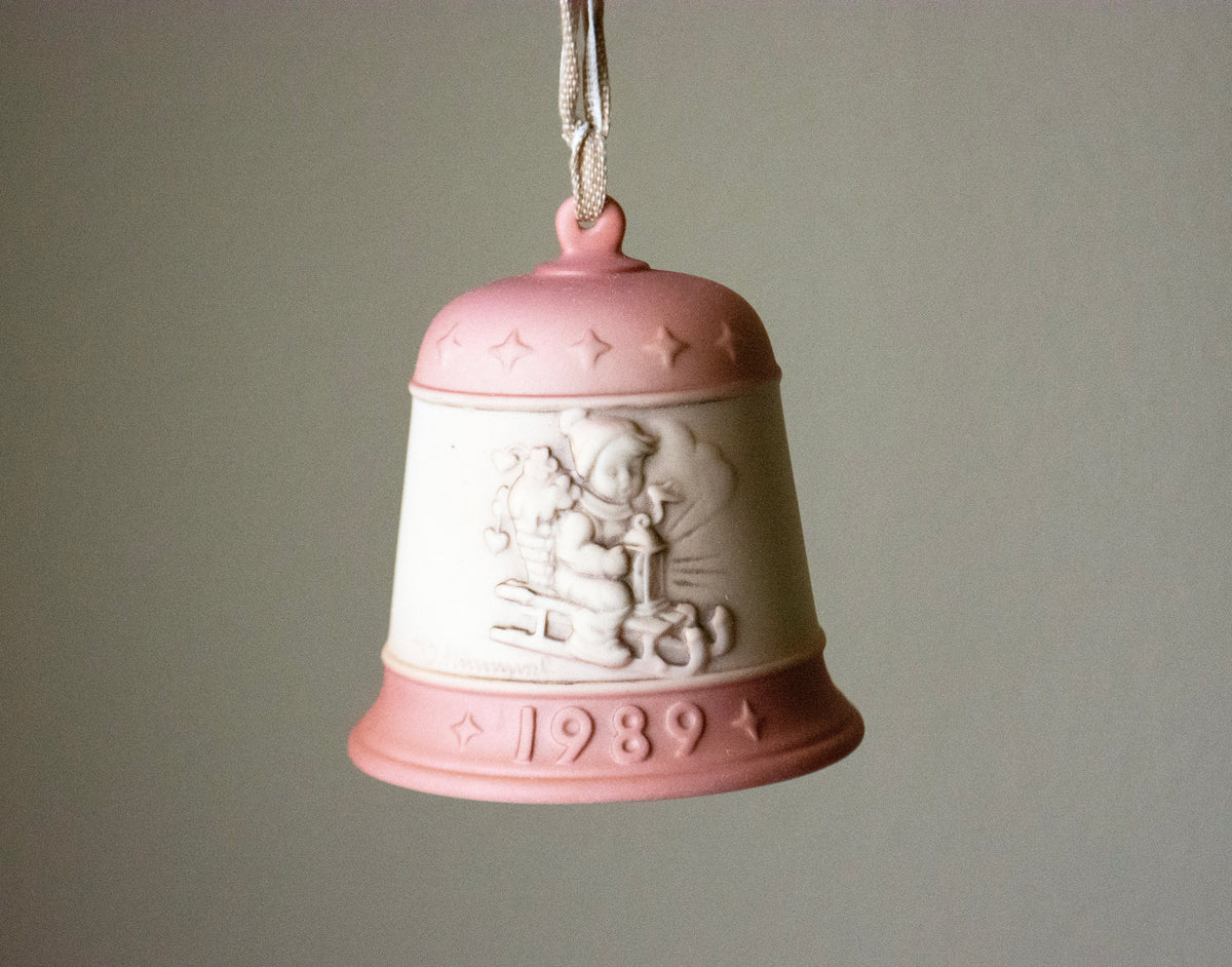 A hanging porcelain Christmas bell ornament in pale pink and off-white, featuring embossed star details, a relief carving of a child on a sleigh, and the year 1989 on the lower rim.