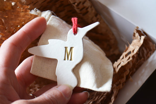 A hand holds a white wooden bird-shaped ornament with a gold letter 'M' and a red hanging string, placed over a small cream fabric pouch inside a gift box with brown paper filler.