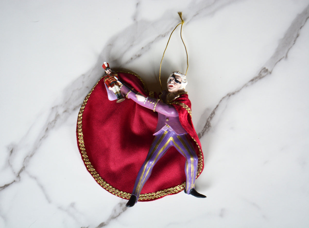 A decorative hanging ornament shaped like a stylized figure in a purple suit with a large red cape and gold trim, posed on a white marble surface.