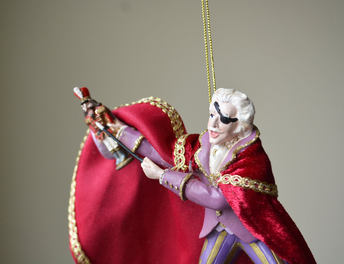 A close-up of a decorative hanging ornament shaped like a theatrical character in a red cape with gold trim, holding a sword, against a plain beige background.