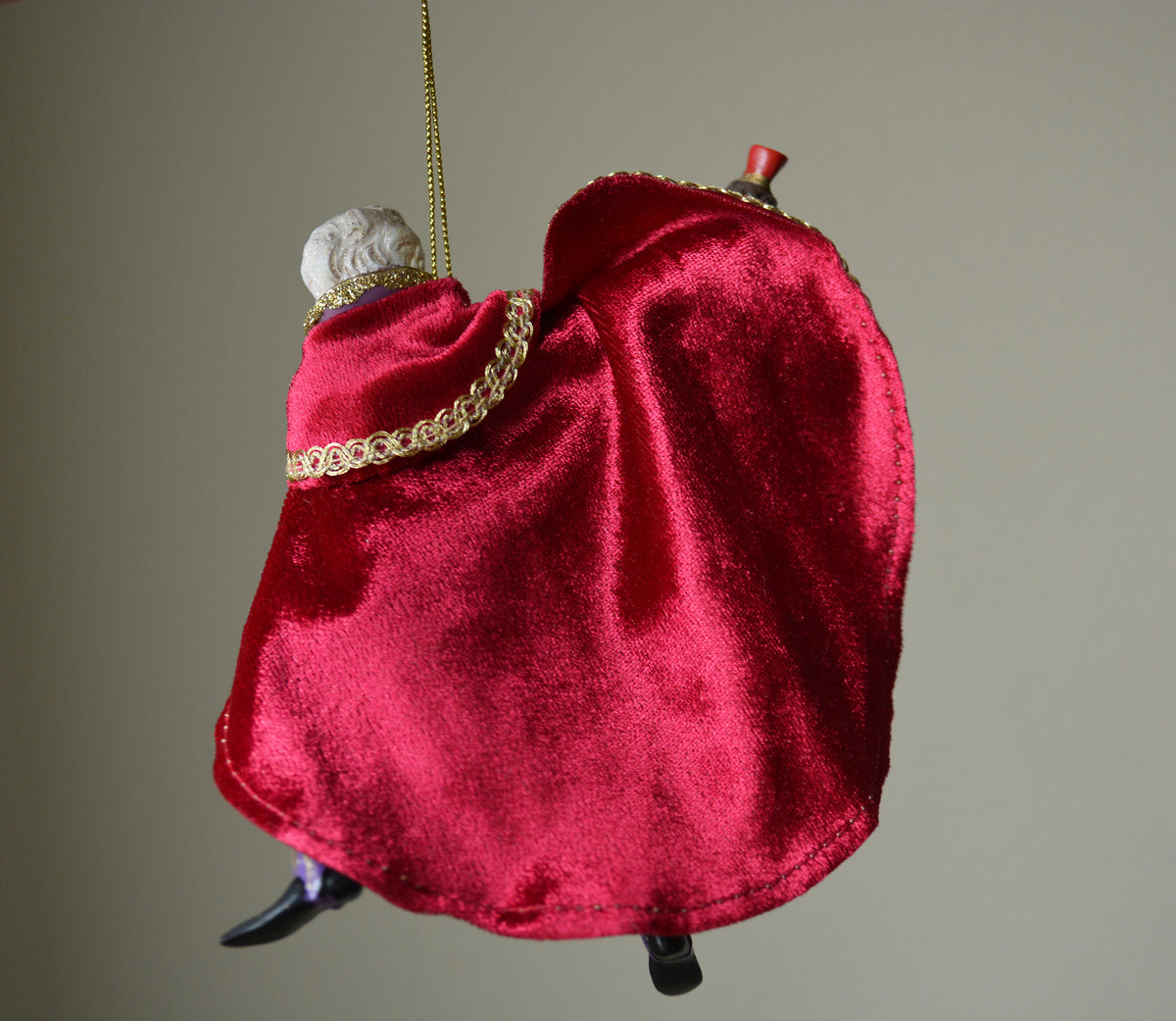 A small hanging ornament shaped like a seated figure draped in bright red velvet fabric, with a gold chain strap, decorative gold trim, a white sculpted head, and black shoes, photographed against a plain warm gray background.