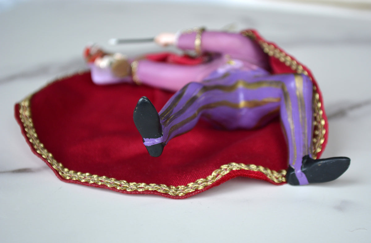 A small decorative figurine of a person lying on a red velvet cushion with gold trim, set on a light marble-like surface. The figure wears purple striped pants, pink sleeves, and black shoes.
