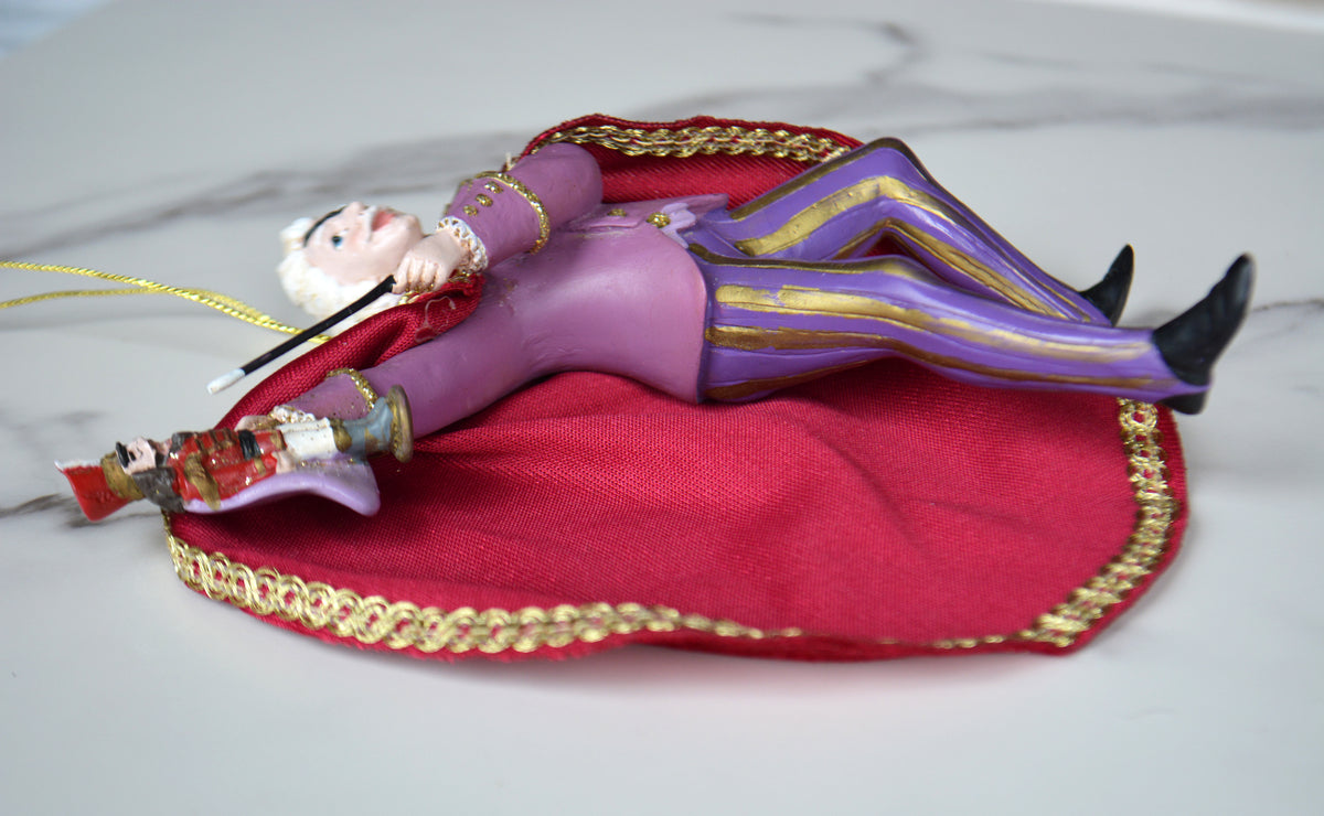 A decorative figurine of a man in a purple suit with gold trim, reclining on a red cape with gold trim, photographed on a light marble-like surface.