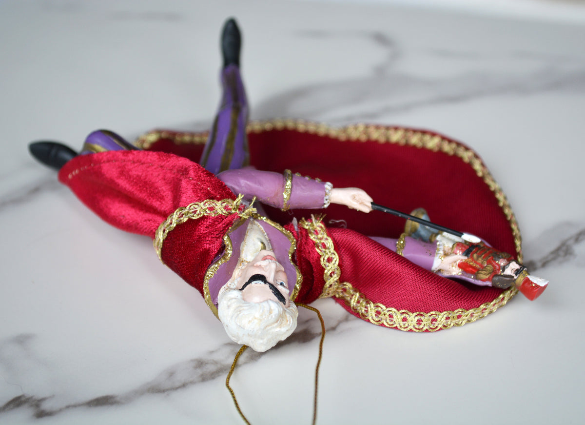 A decorative figurine of a bearded man in a red robe with gold trim and purple sleeves, posed diagonally on a red satin cushion with gold trim, set on a white marble surface.