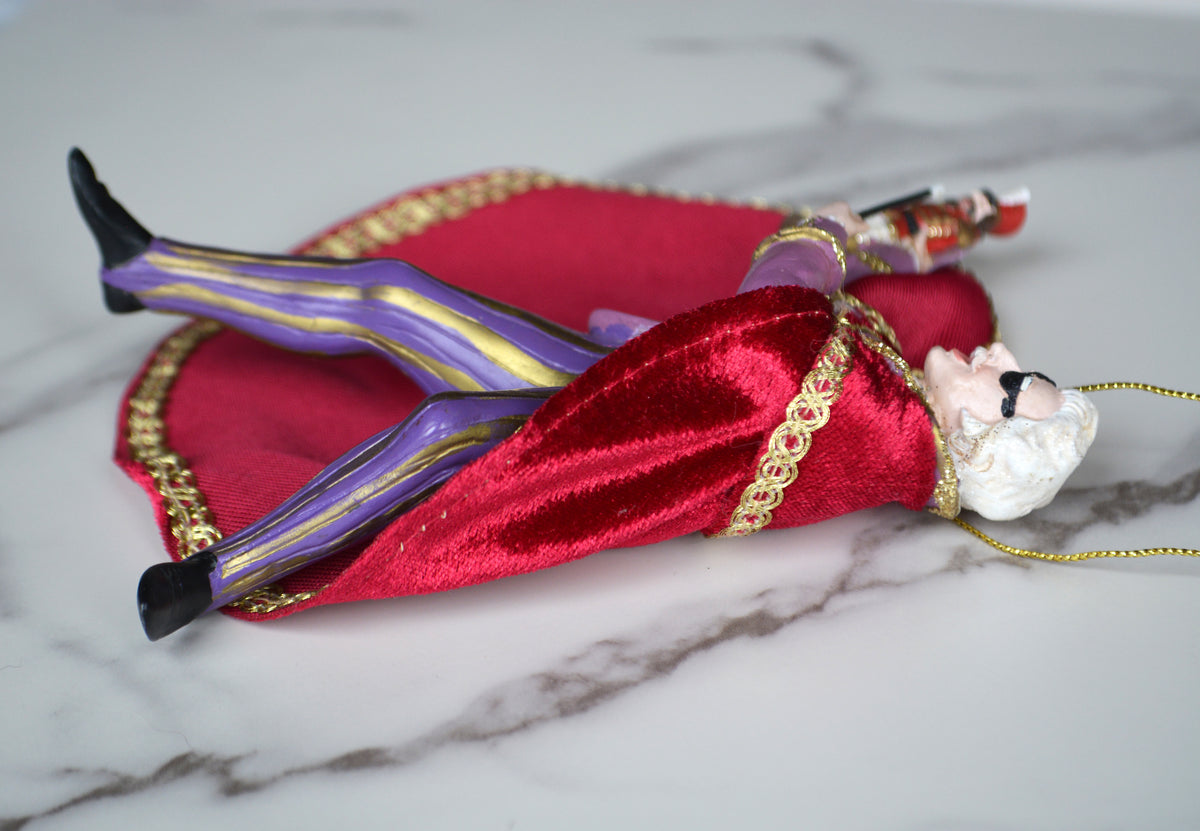 A decorative Santa Claus ornament lying on a white marble surface, wearing a red velvet robe with gold trim, purple and gold striped pants, black shoes, and a white beard.