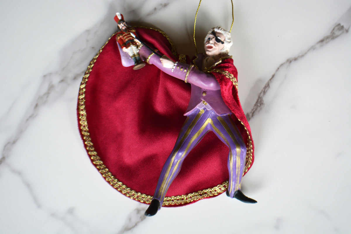 A decorative hanging ornament shaped like a masked performer in a purple outfit with a red cape and gold trim, posed against a large red heart-shaped backdrop on a white marble surface.