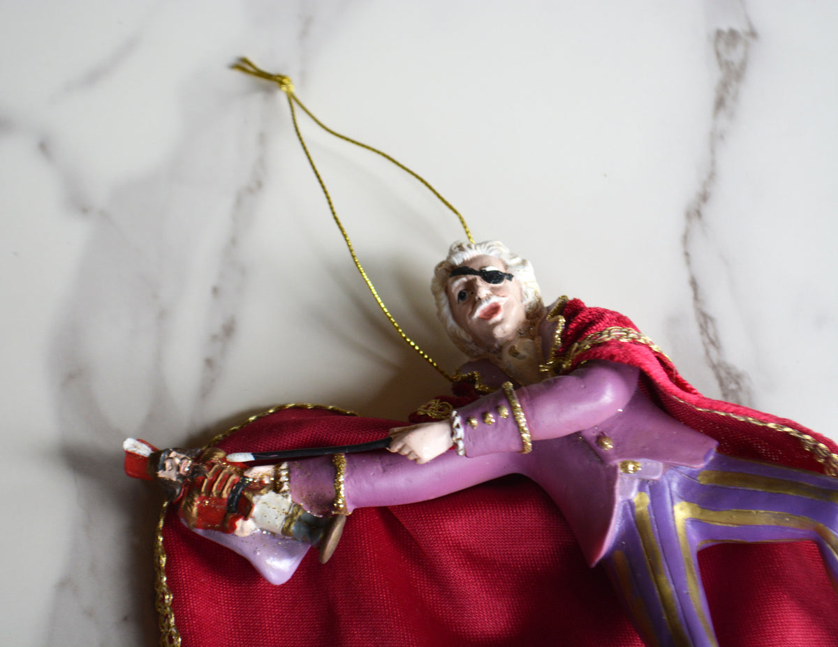A close-up of a decorative figurine or ornament depicting a woman in a purple dress and red cape with gold trim, lying on a white marble surface.