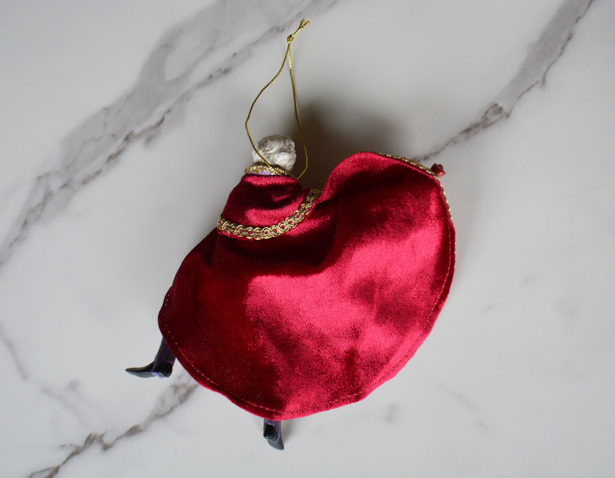 A small red velvet Christmas ornament shaped like a miniature figure in a red dress, with a gold hanging string, gold trim, and small black feet, photographed on a white marble surface.