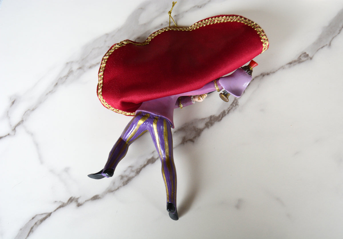 A decorative figurine of a jester-like character wearing a large red velvet hat with gold trim, a purple tunic, and purple-and-gold striped pants, posed on a white marble surface.