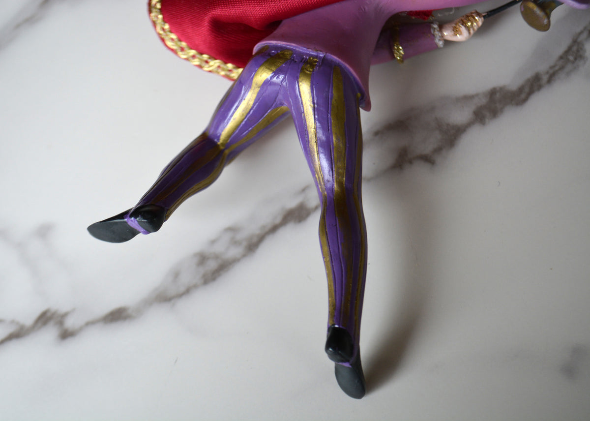 A close-up of a doll's legs wearing purple and gold striped tights, with a pink skirt and red cape, resting on a white marble surface.