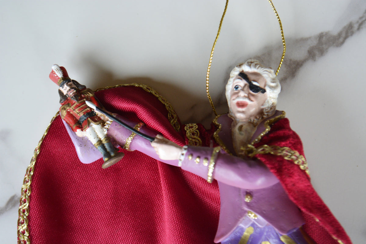 A close-up of a decorative figurine or ornament featuring a woman in a purple outfit with a red draped fabric and gold trim, holding a small soldier figure, on a light marble-like surface.