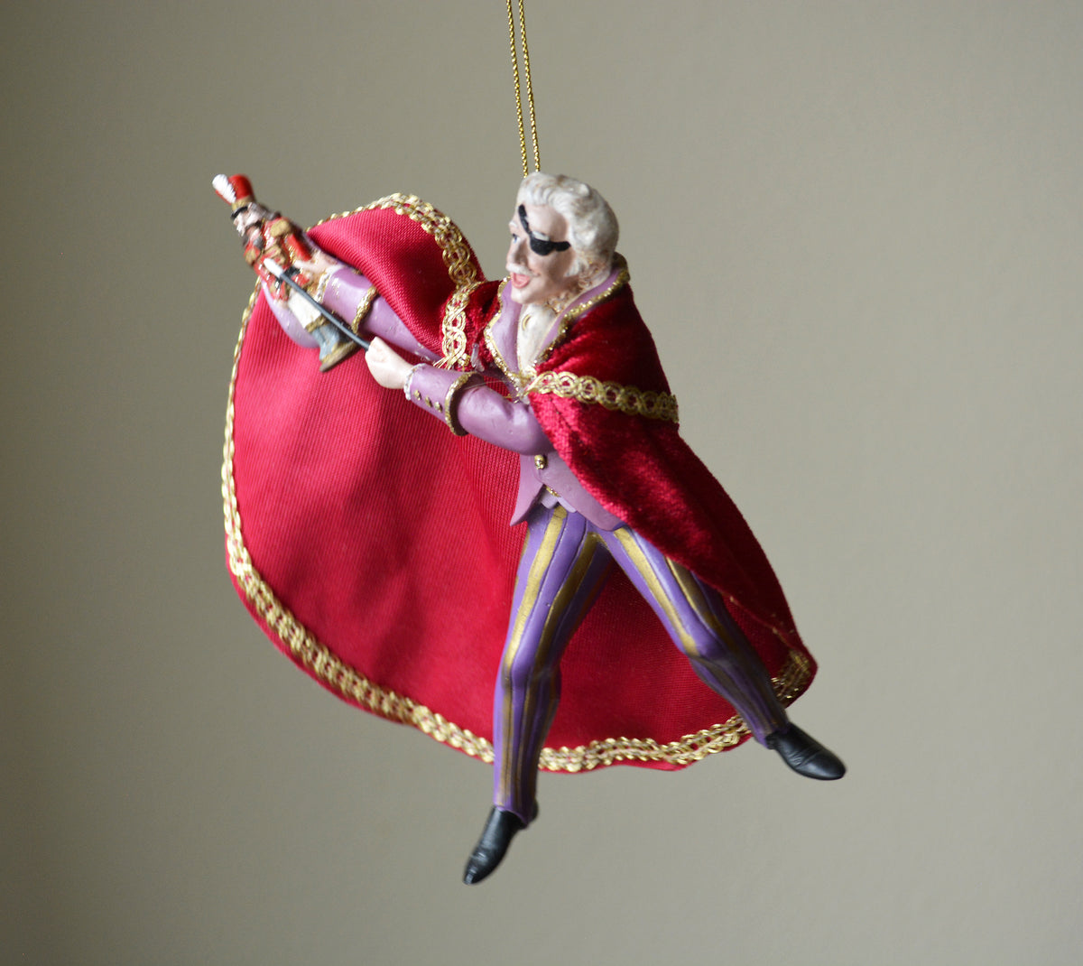 A hanging ornament shaped like a stylized figure in a flowing red cape with gold trim, holding a small nutcracker figure, photographed against a plain warm gray background.
