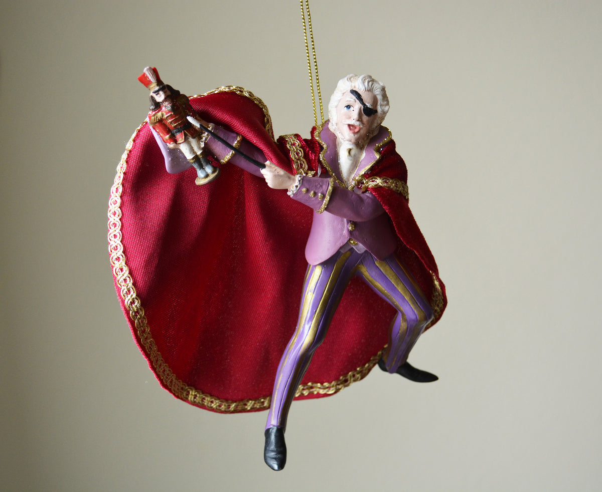 A hanging Christmas ornament shaped like a whimsical character in a purple suit and flowing red cape, holding a nutcracker, photographed against a plain light beige background.