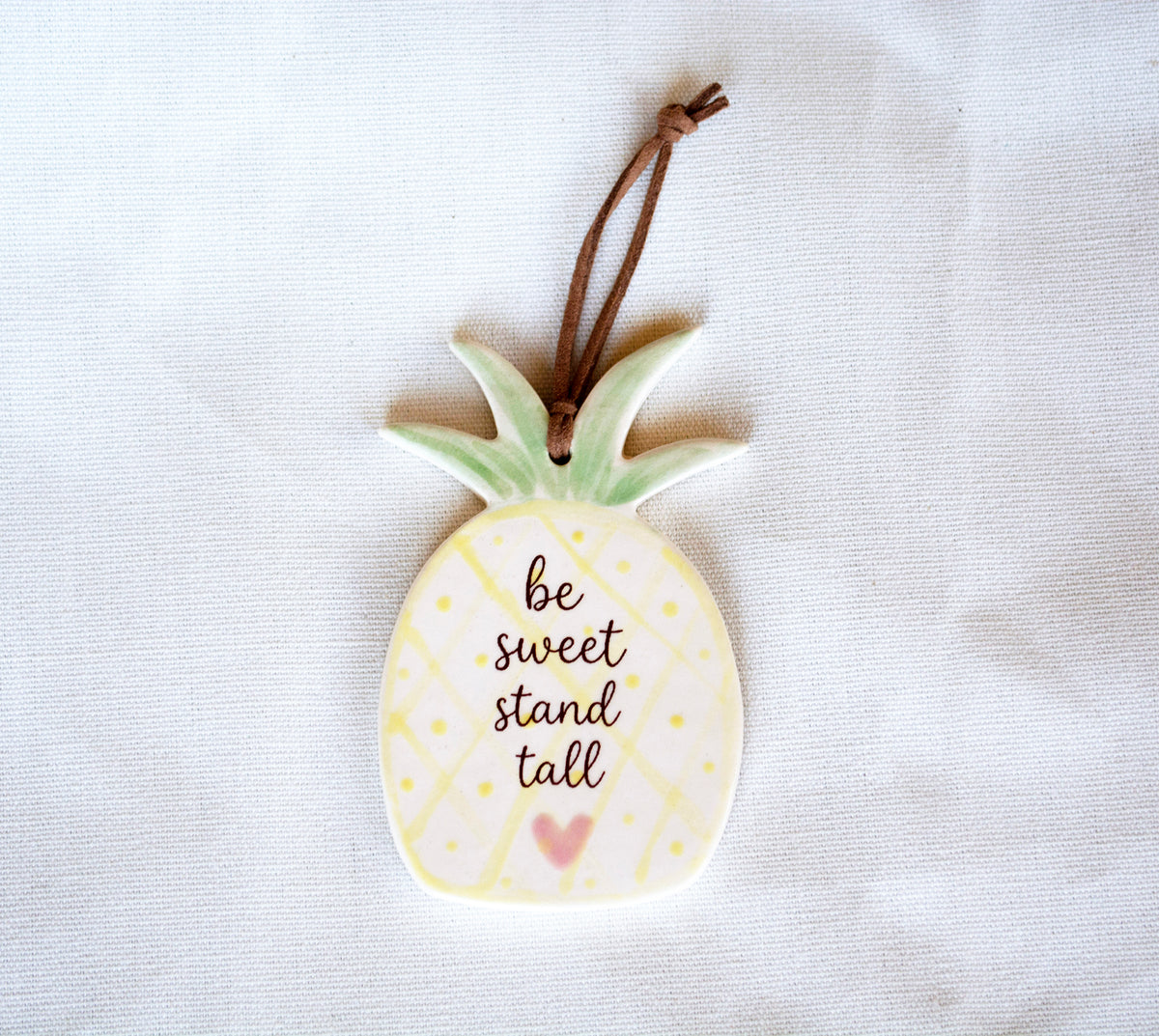 A ceramic pineapple ornament with the phrase 'be sweet stand tall' and a small pink heart, hanging from a brown string against a white fabric background.