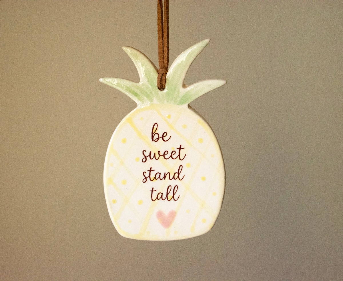 A ceramic pineapple ornament with the phrase 'be sweet stand tall' in brown script, featuring a light green crown, yellow dotted pattern, and a small pink heart, hanging against a plain taupe background.