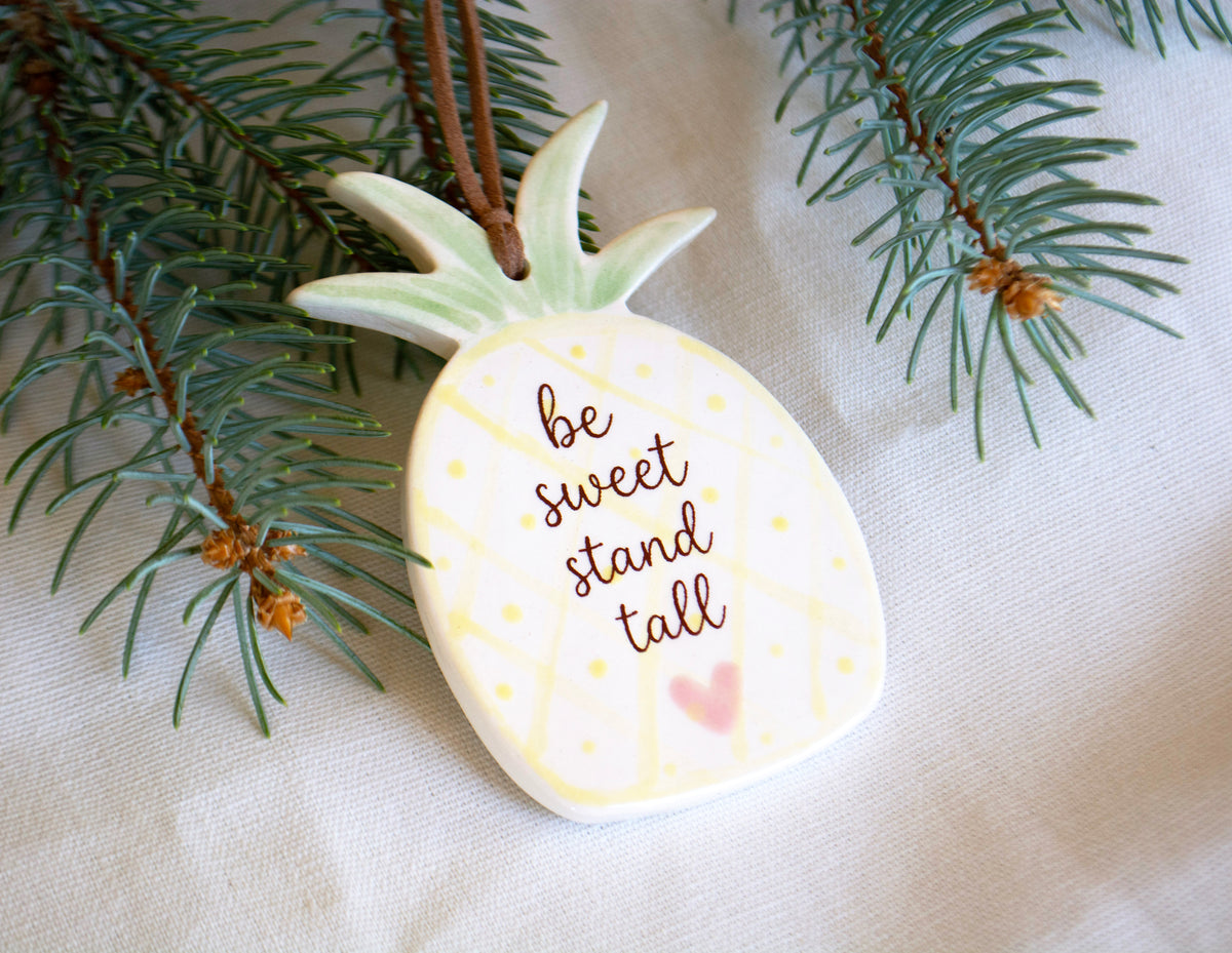 A pineapple-shaped ceramic ornament with the phrase 'be sweet stand tall' in brown script, decorated with yellow dotted lines and a small pink heart. The ornament hangs from a brown string and is styled with green pine branches on a beige fabric background.