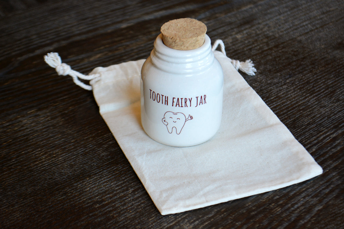 A small white ceramic tooth fairy jar with a cork stopper, printed with 'TOOTH FAIRY JAR' and a tooth illustration, resting on a cream drawstring pouch over a dark wood surface.