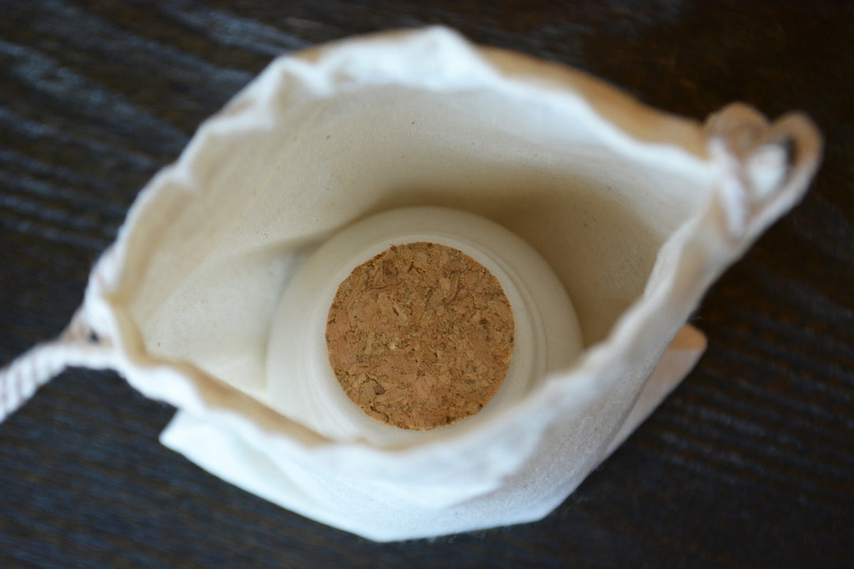 A white fabric drawstring pouch with a cork stopper, shown from a top-down view on a dark wooden surface.