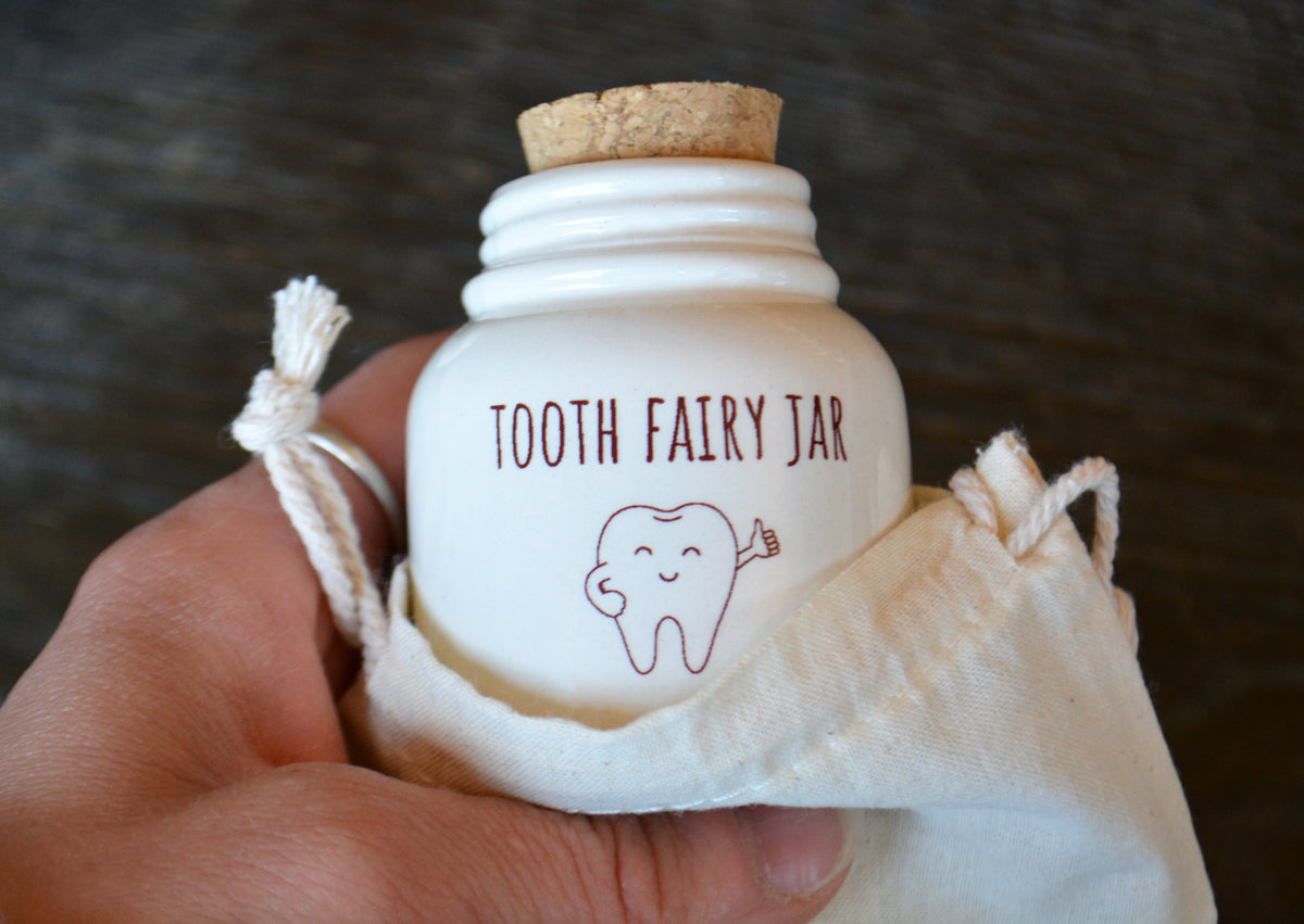 A hand holding a small white jar with a cork stopper and a drawstring fabric pouch, printed with the words 'TOOTH FAIRY JAR' and a tooth illustration.