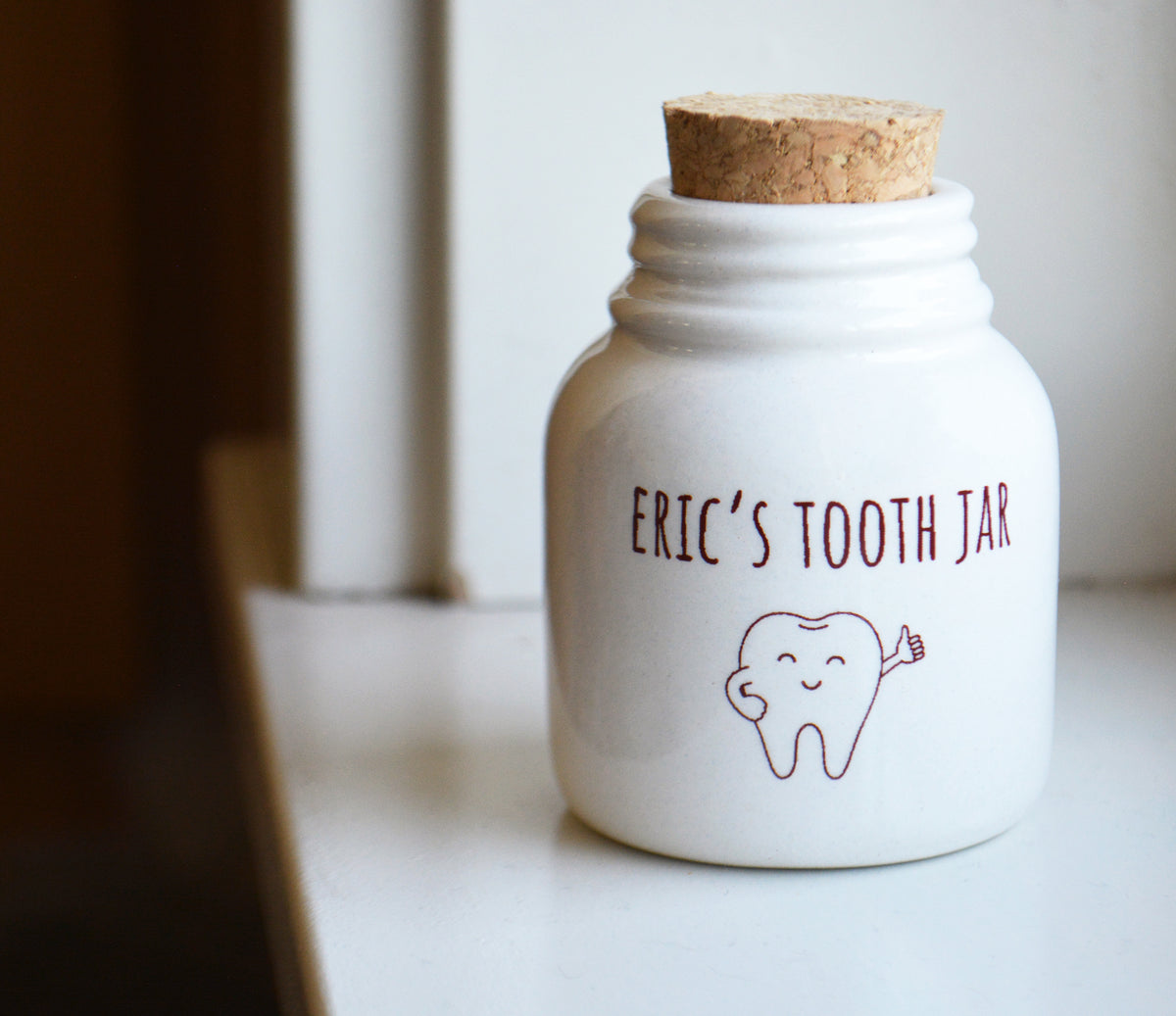 A white ceramic tooth jar with a cork stopper, featuring the text 'ERIC'S TOOTH JAR' and a tooth illustration printed on the front, set on a white surface with a blurred indoor background.