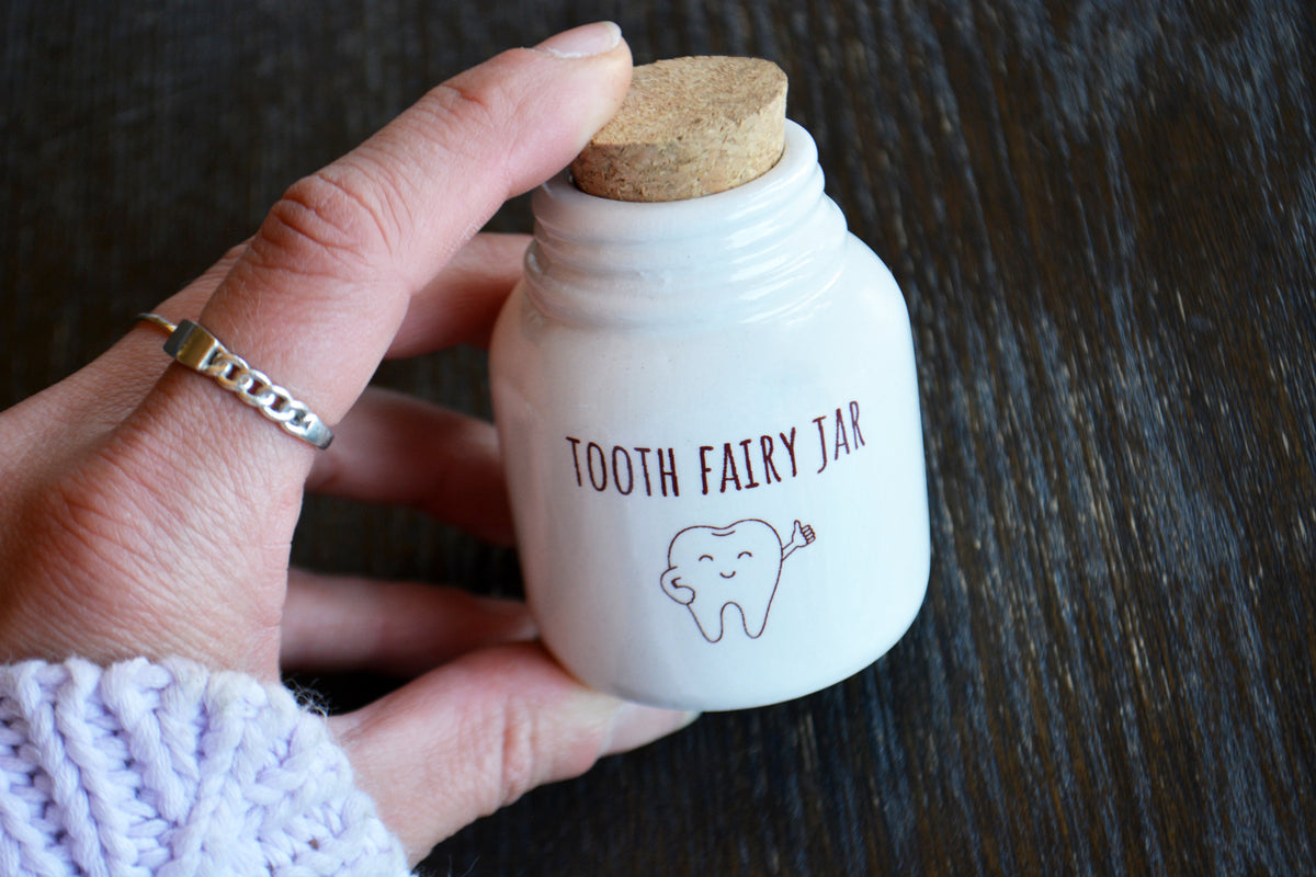 A hand holding a small white ceramic tooth fairy jar with a cork stopper. The jar is printed with the words 'TOOTH FAIRY JAR' and a simple tooth illustration.